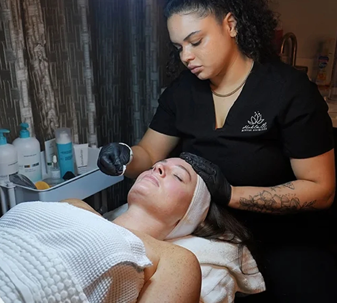 A esthetician wearing a black uniform and black gloves applying a facial treatment to a client lying down in a treatment chair.