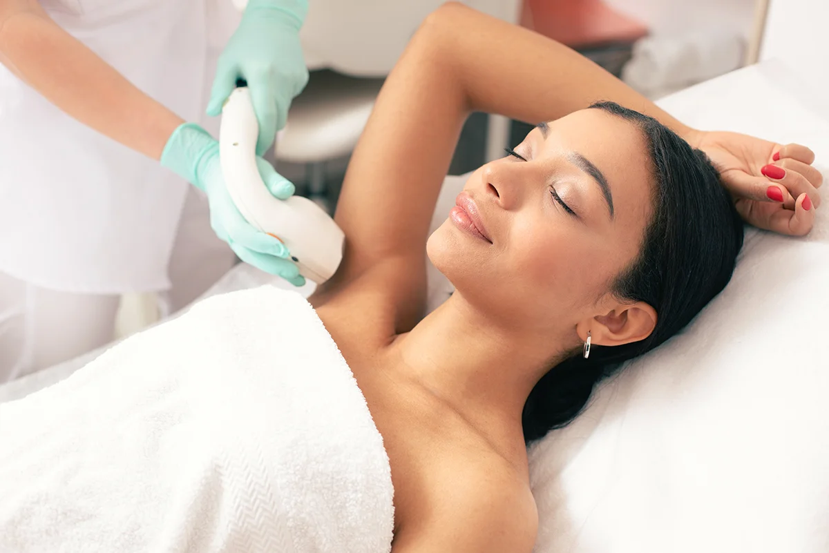 A woman receiving a laser hair reduction treatment on her face while wearing protective eyewear and holding a cooling air hose.