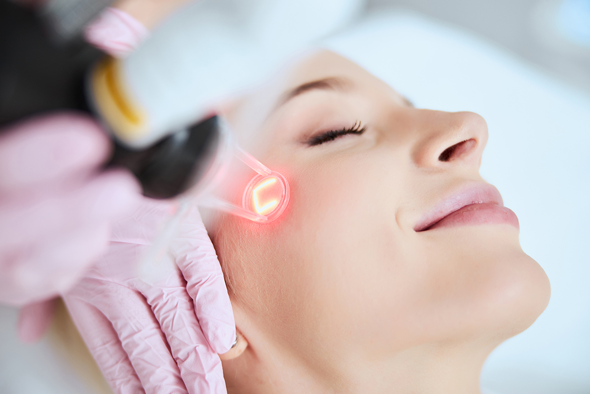 A woman receiving a laser skin treatment on her cheek by a practitioner wearing blue gloves holding a laser handpiece.