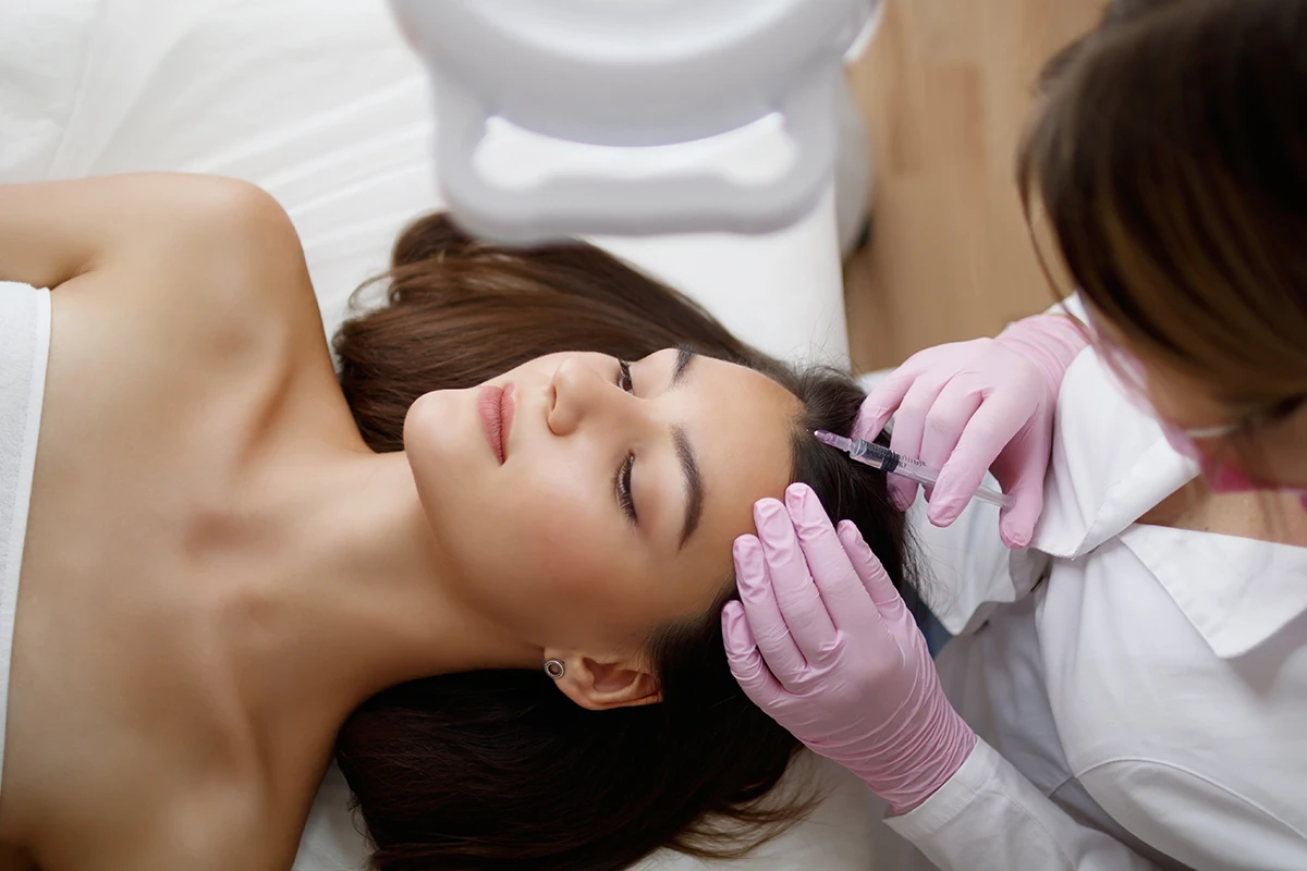 A woman receiving a scalp treatment from an aesthetic provider wearing blue gloves in a clinical setting.