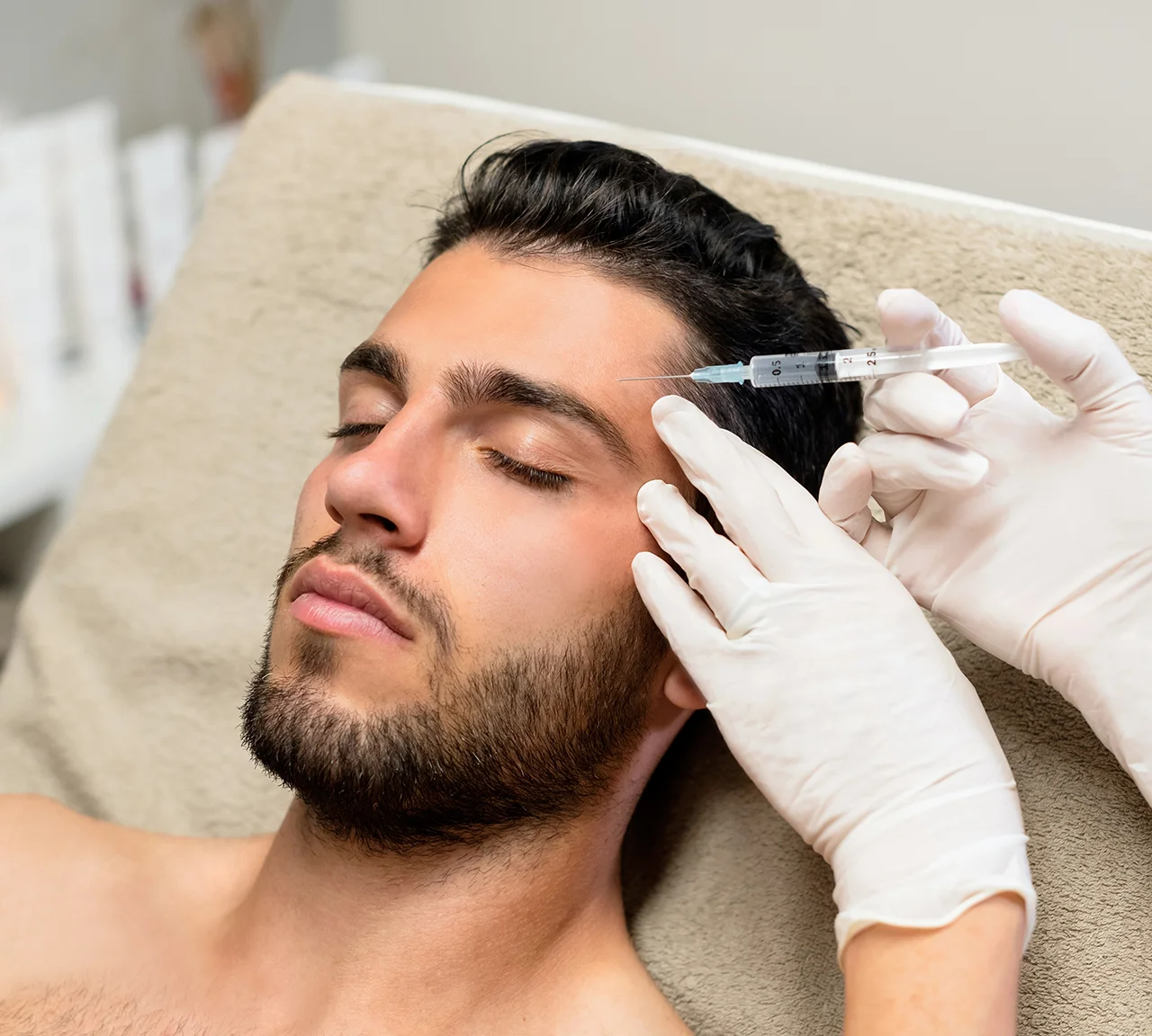 Overhead view of an aesthetic provider in blue latex gloves administering a neurotoxin injection into the forehead of a bearded man.